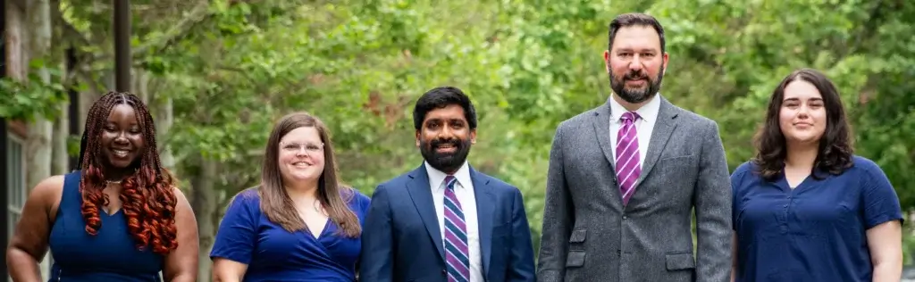 A group of five people standing outdoors, all smiling. They are dressed in formal attire, with trees in the background, conveying a professional and friendly atmosphere.