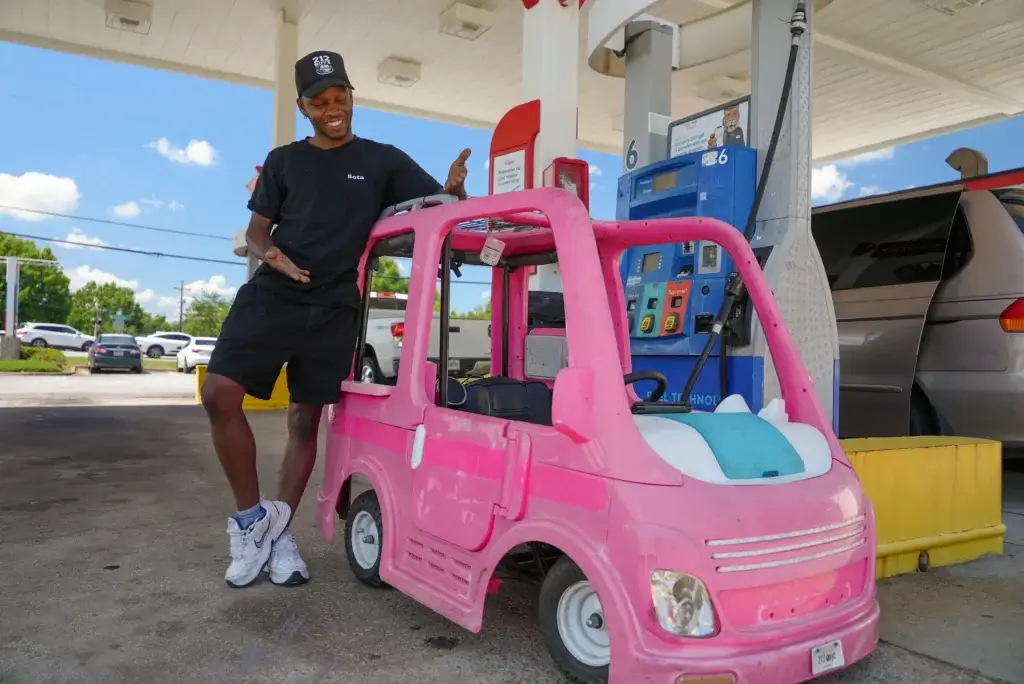 A man stands next to a pink toy car at a gas station pump, smiling with one hand pointing at the car.