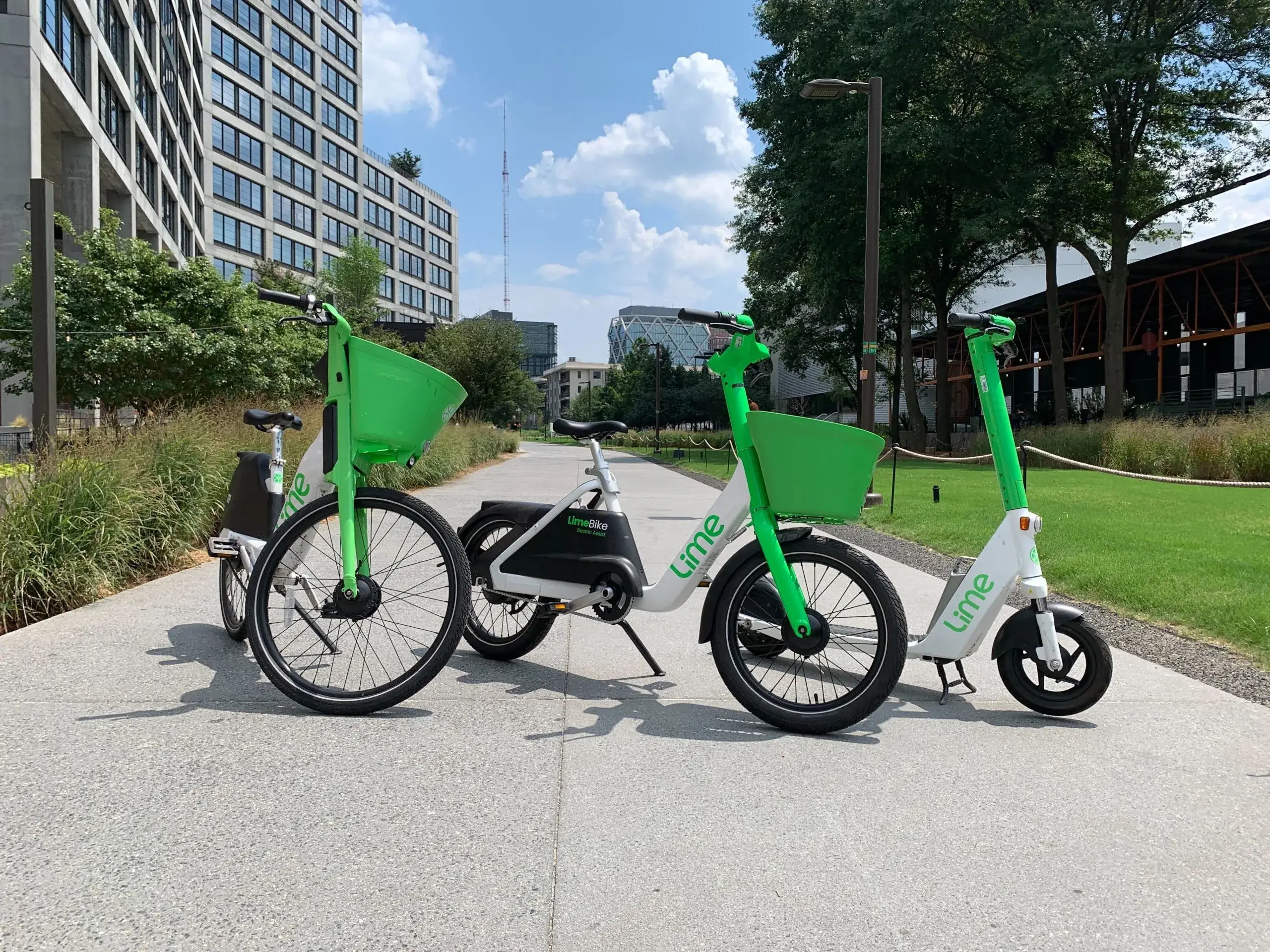 Three Lime-branded electric vehicles—a bike, a moped, and a scooter—are parked on a pathway in an urban outdoor setting on a sunny day.